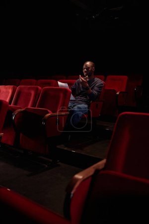 Foto de Fotografía vertical del hombre afroamericano sentado en la audiencia y leyendo líneas antes del espectáculo en el espacio de copia del teatro vacío - Imagen libre de derechos
