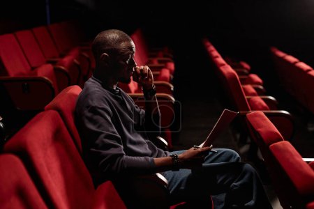Foto de Retrato de vista lateral del hombre afroamericano sentado en audiencia y leyendo guion en espacio de copia de teatro vacío - Imagen libre de derechos