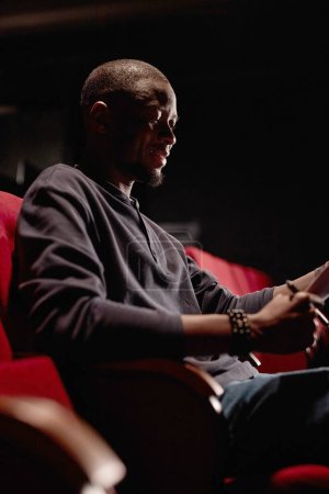 Foto de Retrato vertical del hombre afroamericano sentado en audiencia con poca luz y guion de lectura - Imagen libre de derechos