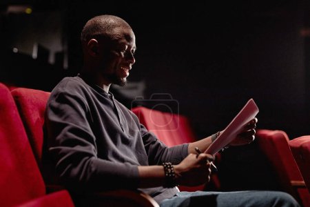 Foto de Retrato de vista lateral del hombre negro sonriente sentado en la audiencia y leyendo guion con poca luz en el espacio de copia del teatro - Imagen libre de derechos