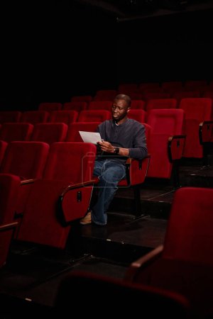 Foto de Retrato vertical mínimo del hombre afroamericano como director de teatro leyendo guion sentado solo en audiencia - Imagen libre de derechos