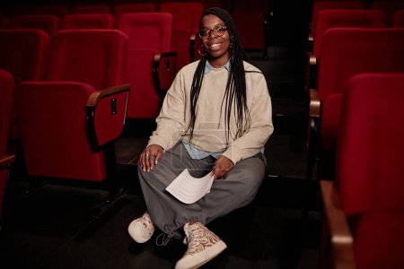 Foto de Retrato de larga duración de una mujer afroamericana sonriente sentada en el suelo en el teatro y mirando el espacio fotográfico - Imagen libre de derechos