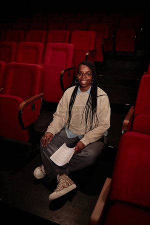 Téléchargez les photos : Portrait vertical complet de la femme afro-américaine adulte en tant qu'interprète féminine assise sur le sol dans le théâtre avec les bras croisés et regardant la caméra - en image libre de droit