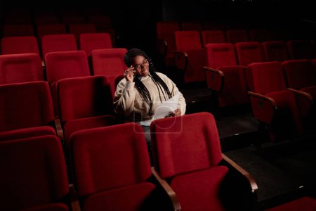 Foto de Retrato mínimo de una mujer afroamericana sonriente leyendo líneas mientras está sentada en una audiencia vacía en el espacio de copia del teatro - Imagen libre de derechos