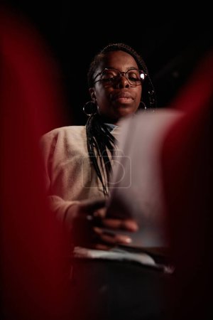 Foto de Retrato vertical de bajo ángulo de la mujer afroamericana leyendo guion sentado en audiencia en el teatro y preparándose para el espectáculo - Imagen libre de derechos