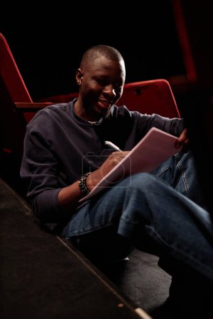 Foto de Retrato vertical del intérprete afroamericano escribiendo guion y ensayando líneas sentadas en el suelo en el teatro - Imagen libre de derechos