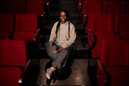 Téléchargez les photos : Portrait complet de la femme adulte afro-américaine souriant à la caméra tout en étant assis sur le sol dans l'espace de copie de théâtre - en image libre de droit