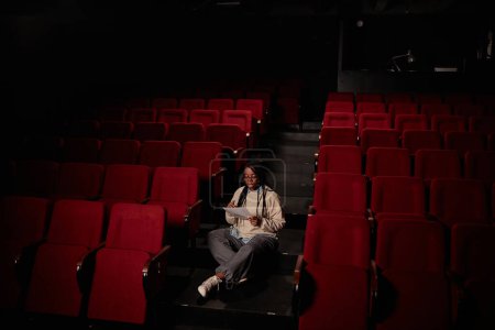 Téléchargez les photos : Vue grand angle minime à l'artiste afro-américaine féminine répétant des lignes assises seules dans le public du théâtre avec un espace de copie faible lumière - en image libre de droit
