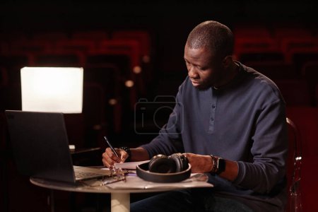 Porträt eines erwachsenen schwarzen Mannes als Theaterregisseur, der sich Notizen macht, am Tisch sitzend mit Laptop-Kopierplatz