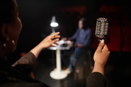 Close up of female hands holding retro microphone while performing on stage with theater director watching copy space