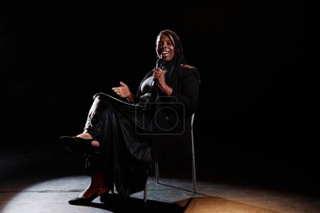 Minimal full length portrait of smiling Black woman performing comedy show while sitting in chair on stage and speaking to microphone copy space