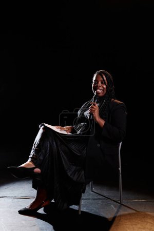 Vertical full length portrait of smiling young woman speaking to microphone on stage while sitting in chair with spotlight