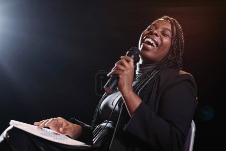 Portrait of Black woman laughing on stage and holding microphone while performing in comedy show copy space