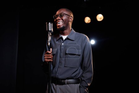 Retrato de la cintura hacia arriba del joven negro sonriente hablando con el micrófono en el escenario mientras actúa en el espectáculo de comedia