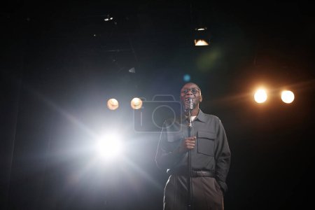 Wide angle shot of African American man speaking to microphone standing on stage with spotlight and smiling copy space