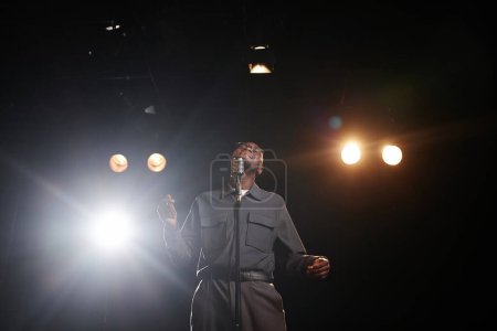 Low angle shot of young Black man performing on stage in theater and dancing with microphone copy space