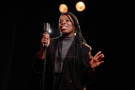 Téléchargez les photos : Portrait taille haute d'une élégante femme noire se produisant sur scène avec microphone sur fond sombre espace de copie - en image libre de droit