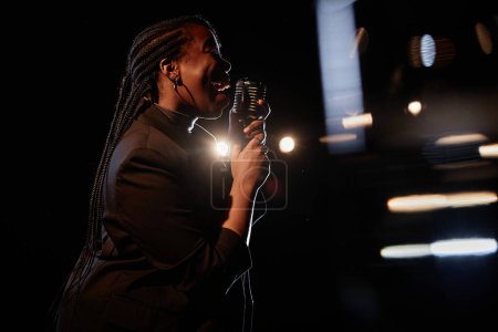Side view portrait of African American woman singing to microphone while performing on stage in dark