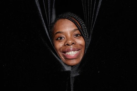 Primer plano de la sonriente mujer afroamericana asomándose desde las cortinas en el escenario del teatro y mirando a la cámara