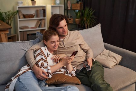 Téléchargez les photos : Portrait de jeune couple regardant la télévision à la maison ensemble assis sur un canapé confortable - en image libre de droit