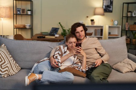 Front view portrait of young couple watching TV at home together sitting on comfortable sofa and cuddling