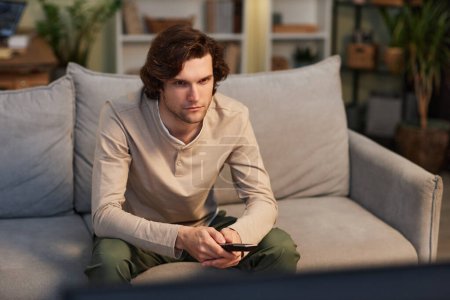 Téléchargez les photos : Portrait de jeune homme regardant la télévision à la maison assis sur le canapé dans un intérieur vert confortable, espace de copie - en image libre de droit