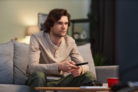 Téléchargez les photos : Portrait de jeune homme regardant la télévision à la maison assis sur le canapé intérieur, espace de copie - en image libre de droit