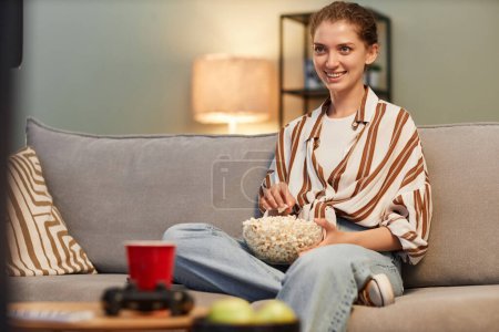 Foto de Retrato de una joven sonriente viendo la televisión en casa y comiendo palomitas de maíz, espacio para copiar - Imagen libre de derechos