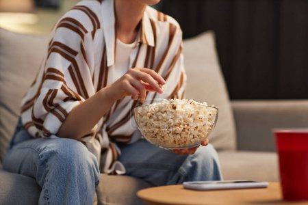 Téléchargez les photos : Gros plan de jeune femme méconnaissable regardant la télévision à la maison et mangeant du pop-corn, espace de copie - en image libre de droit