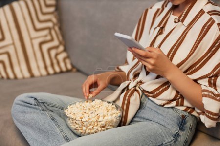 Closeup of unrecognizable young woman relaxing at home with smartphone and eating popcorn, copy space
