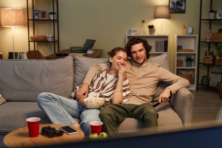 Retrato de vista frontal de feliz pareja joven viendo películas en casa y comiendo palomitas de maíz sentado en un cómodo sofá, espacio para copiar