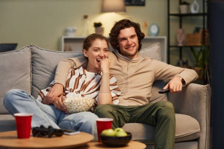 Téléchargez les photos : Portrait de jeune couple heureux regardant des films à la maison et mangeant du pop-corn assis sur le canapé ensemble - en image libre de droit