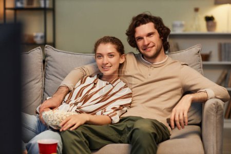 Photo for Portrait of smiling couple watching movies at home and eating popcorn sitting on couch together - Royalty Free Image