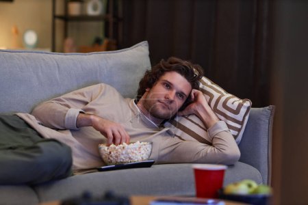 Foto de Retrato de un hombre joven viendo Tv en casa y comiendo palomitas de maíz mientras está acostado en el sofá, espacio para copiar - Imagen libre de derechos
