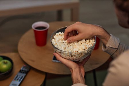 Photo for Closeup of young man eating popcorn from glass bowl while watching TV or movies at home, copy space - Royalty Free Image