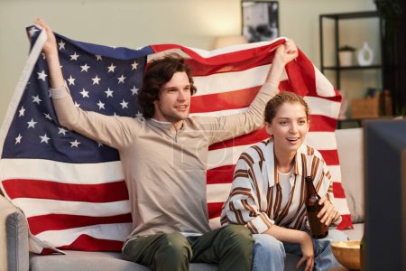 Téléchargez les photos : Portrait de jeune couple avec drapeau américain acclamant tout en regardant match de sport à la maison - en image libre de droit