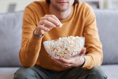 Closeup of young man holding glass bowl with popcorn while watching TV at home, copy space