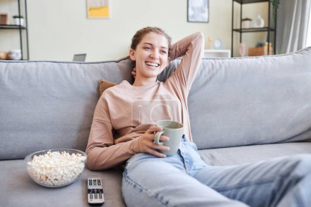 Portrait of relaxed young woman watching TV at home and laughing, copy space