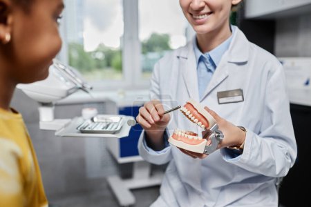 Photo for Closeup of friendly woman dentist holding tooth model and explaining dental hygiene to little girl, copy space - Royalty Free Image
