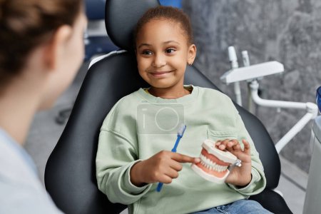 Téléchargez les photos : Portrait de petite fille noire dans une chaise dentaire tenant le modèle de dent et souriant regardant joyeusement le dentiste féminin, espace de copie - en image libre de droit