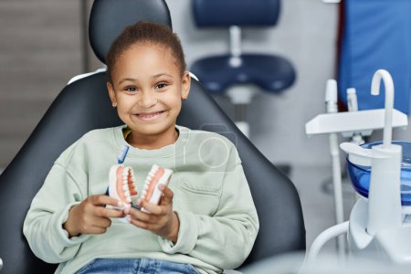Photo for Front view portrait of black little girl in dental chair smiling at camera happily and holding tooth model, copy space - Royalty Free Image