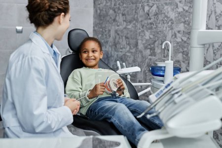 Photo for Portrait of smiling little girl sitting in dental chair and listening to female dentist in consultation on tooth health, copy space - Royalty Free Image