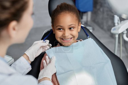 Photo for Portrait of smiling little girl in dental chair with nurse preparing her for teeth check up, copy space - Royalty Free Image