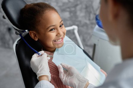 Téléchargez les photos : Portrait de mignonne fille noire dans une chaise dentaire avec infirmière ou dentiste la préparant à l'examen des dents, espace de copie - en image libre de droit