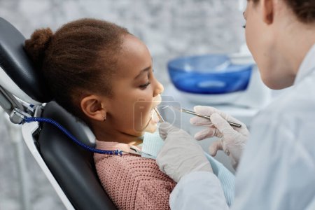 Téléchargez les photos : Portrait latéral de la petite fille noire dans une chaise dentaire pendant le contrôle des dents avec un pédiatre dentiste, espace de copie - en image libre de droit