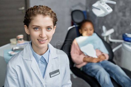 Photo for Portrait of young woman as dentist smiling at camera in clinic with little kid in background, copy space - Royalty Free Image