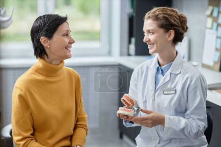 Photo for Side view portrait of smiling female dentist holding tooth model and talking to mature woman in clinic - Royalty Free Image