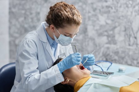 Photo for Side view portrait of young female dentist wearing mask while working with female patient in dental clinic, copy space - Royalty Free Image