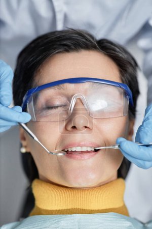 Téléchargez les photos : Gros plan de femme mature allongée sur une chaise dentaire avec les yeux fermés pendant l'examen des dents - en image libre de droit