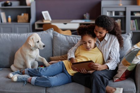 Téléchargez les photos : Portrait pleine longueur de mère et fille aimante en utilisant une tablette numérique avec chien mignon tout en se relaxant sur le canapé à la maison - en image libre de droit